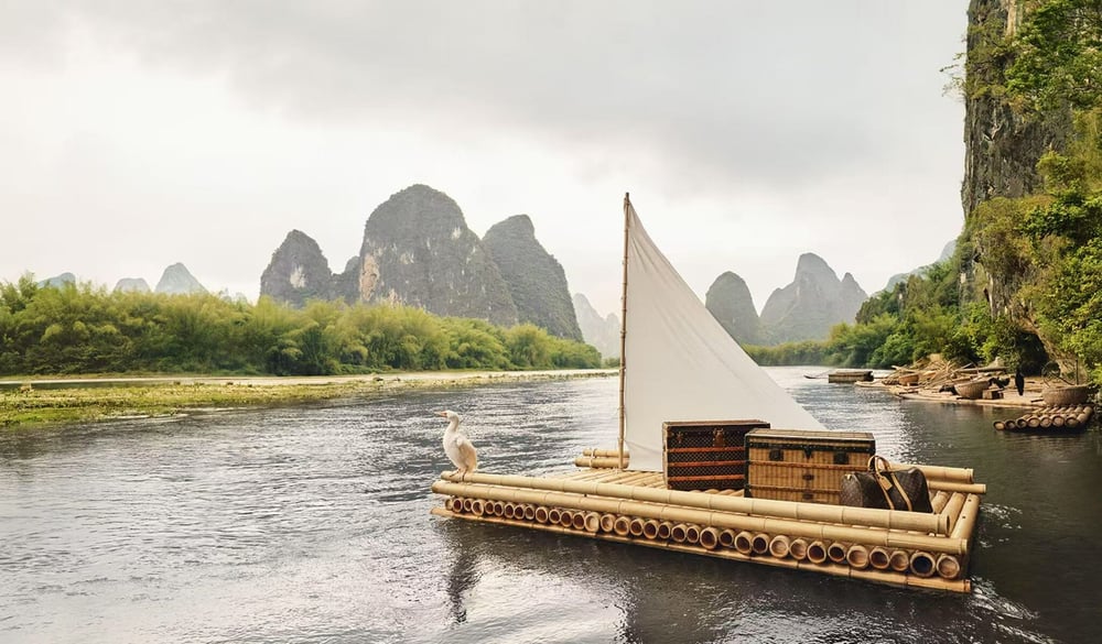 Louis Vuitton’s new travel campaign captures the serene beauty of the Lijiang River, blending iconic trunks and the Soft Keepall with traditional bamboo rafts to reflect a harmonious dialogue between heritage and nature.